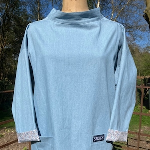 May include: A light blue denim smock with a patch pocket and a floral print on the cuffs. The smock has a high neck and long sleeves. The label on the pocket reads "SMOCK".