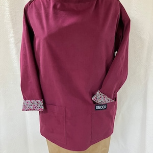 May include: A burgundy smock with long sleeves and a patch pocket on the front. The sleeves and neckline are lined with a floral print fabric. The smock has a label that says "SMOCK" on the front.