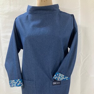May include: A blue denim smock with a high neck and long sleeves. The sleeves have white and blue floral patterned cuffs. The smock has a single pocket on the front with the word "SMOCK" printed on a white label.