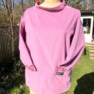 May include: A pink smock with long sleeves and a patch pocket on the front. The smock has a floral print on the inside of the collar and cuffs. The word "SMOCK" is printed on the pocket.