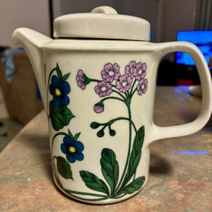 Op de afbeelding: Witte theepot van keramiek met een bloemenmotief met roze, blauwe en groene bloemen. De theepot heeft een deksel en een handvat.