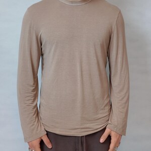 Könnte beinhalten: Ein langärmeliges Shirt mit Rundhalsausschnitt in einem einfarbigen Hellbraun. Das Kleidungsstück verfügt über einen einfachen runden Halsausschnitt und lange Ärmel, was für ein schlichtes und neutrales Erscheinungsbild sorgt. Der Stoff ist glatt.