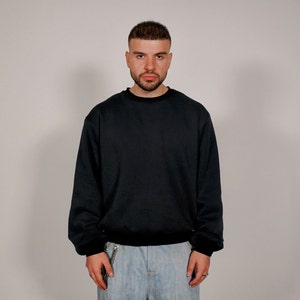 Könnte beinhalten: Ein Mann trägt einen schwarzen Pullover und eine hellblaue Jeans. Er hat kurze, dunkle Haare und einen Bart.