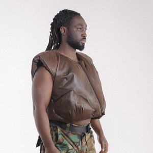 Puede incluir: Un chaleco de cuero marrón con cierre de cremallera y diseño acolchado. El chaleco se lleva sobre una camisa de estampado de camuflaje.