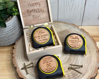 May include: Three black and yellow tape measures with personalized wooden circular inserts, one inside a wooden box engraved with "Happy Birthday Ben" and two placed outside, accompanied by four metal screws on a natural wood slice base.