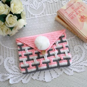 Può includere: Una pochette all'uncinetto rosa, bianca e grigia fatta a mano con un motivo a cesta intrecciata. La pochette presenta una chiusura con pompon bianco e un bottone di legno. Il design include una patta. Un accessorio elegante.