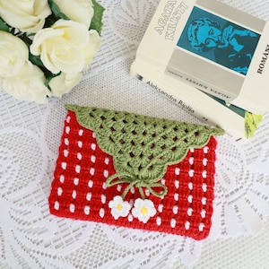 Op de afbeelding: Een rode en witte gehaakte clutch in de vorm van een aardbei met groene gehaakte bladeren en witte gehaakte bloemen. De clutch ligt op een wit kantkleed.