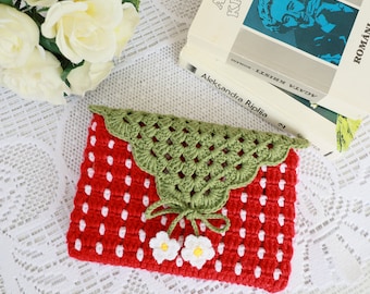 May include: A red and white crocheted strawberry-shaped clutch with green crocheted leaves and white crocheted flowers. The clutch is sitting on a white lace tablecloth.