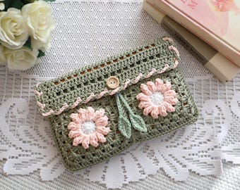 May include: A handmade crochet clutch in olive green, featuring two pink and white daisy appliqués. The clutch has a button closure and is trimmed with pink yarn. The design includes two green leaves. The clutch is on a white lace surface.