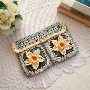 Può includere: Pochette all'uncinetto fatta a mano nei toni del verde, crema e giallo, con un motivo floreale. La pochette ha una chiusura con bottone ed è realizzata con un motivo a granny square. Il design complessivo è fatto a mano e ha un'estetica vintage.