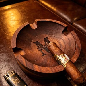 May include: A wooden ashtray with a cigar resting in it, and a lighter. The ashtray is dark brown with the word "MOREL" and the letter "M" engraved on it. The lighter is black with gold trim and the letter "M".