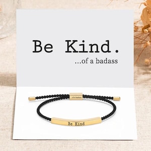 Op de afbeelding: Zwart koord armband met een gouden staaf bedel met de tekst "Be Kind...of a badass".