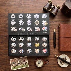 Può includere: Una collezione di mini palloni della Coppa del Mondo 1970-2026 in una scatola nera contenente quindici piccoli palloni da calcio, ognuno rappresentante un anno di torneo. Il coperchio mostra illustrazioni dei palloni con anni e paesi ospitanti.