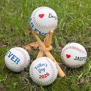 Może przedstawiać: Białe piłki do baseballu z czerwonymi szwami i niebieskim tekstem. Jedna piłka znajduje się na drewnianym tee. Piłki do baseballu mają tekst "Forever", "Benjamin", "Father's Day 2026" i "Jasper". Każda piłka ma czerwone serce.