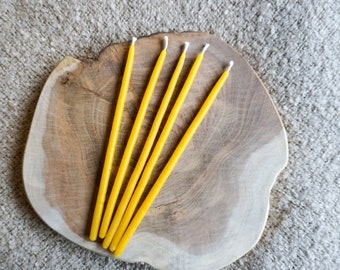 May include: Five yellow beeswax candles on a wooden surface. The candles are arranged in a fan shape.