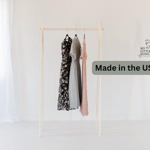 May include: A wooden clothing rack with three dresses hanging on it. The dresses are black, gray, and pink. The rack is made in the USA.  The rack is standing on a white floor.
