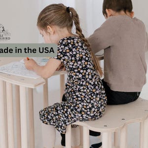May include: Two children sit at a wooden table with rounded edges. The girl on the left is wearing a black and white floral dress and is coloring in a coloring book. The boy on the right is wearing a brown sweater and black pants. The table is made of wood and has a natural finish. The text "Made in the USA" is visible in the image.