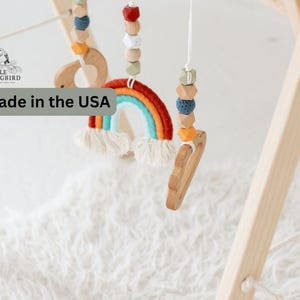 May include: A wooden baby play gym with a rainbow-colored felt rainbow hanging from it. The gym is made of natural wood and has a white fuzzy rug underneath it. The text "Made in the USA" is visible in the image.