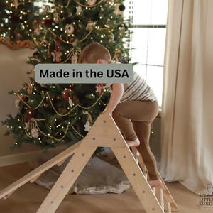 May include: A natural wood climbing structure with a triangular frame and a ramp. The structure is designed for children's play and is shown indoors. The text "Made in the USA" is visible.
