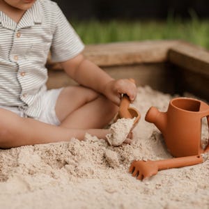 May include: A child plays in a sandbox with a set of terracotta-colored sand toys. The set includes a watering can, a rake, and a scoop. The child is wearing a striped shirt and white shorts. The sandbox is made of wood.
