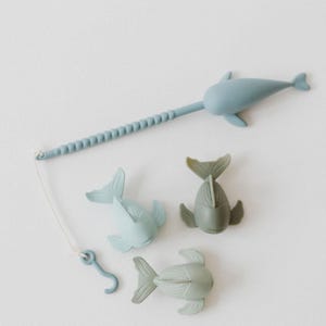 May include: A light blue toy fishing rod with a fish-shaped handle and a hook, along with three fish toys in shades of blue and green. The toys are designed for children.