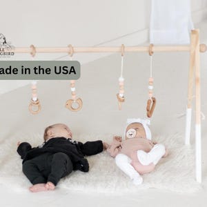 May include: A wooden baby play gym with a white and natural wood finish. The gym has a wooden bar with hanging toys, including a wooden ring, a wooden bird, and a beaded string. Two babies are playing on a white fluffy rug under the gym. The text "Made in the USA" is visible in the image.