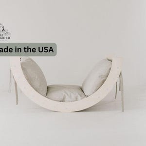 May include: A white wooden rocking chair with two beige cushions. The chair is shaped like a crescent moon and has a small logo that says "My Little Songbird" on the back. The chair is made in the USA.