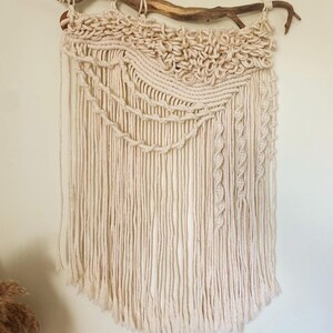 May include: Cream-colored macrame wall hanging. The textile art piece features a knotted design with a textured top section, cascading fringe, and decorative rope details. It is suspended from a natural wooden branch, adding a rustic touch to the decor.