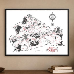 May include: A framed black and white map of The County of Surrey with red text accents. The map features detailed illustrations of forests, a compass rose, and a scale bar. The title The County of Surrey is printed in red in the bottom right corner.