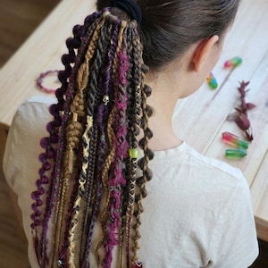 May include: A ponytail with a variety of dreadlocks in shades of purple, brown, and gold. The dreadlocks are adorned with beads and small charms, including a skull. The hair is styled with a black hair tie.