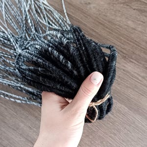 Op de afbeelding: Een bundel zwarte en grijze dreadlocks vastgebonden met touw. De dreadlocks zijn een mix van zwarte en grijze strengen, met een getextureerd uiterlijk. De achtergrond is een houten oppervlak.