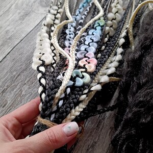 May include: Close-up of a bundle of braided hair extensions in various colors. The braids include black, white, and gray strands, with accents of pastel blue, pink, and green. The bundle is tied with twine.