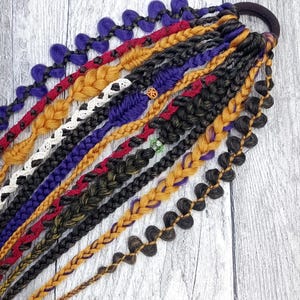 May include: A selection of vibrant braided hair extensions in shades of purple, red, gold, black, and brown. The braids are secured to a black hair tie, displaying a variety of textures and designs. These extensions are designed to add length and volume to hair.