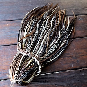 May include: A collection of brown, black, and white synthetic dreadlocks, bound with a cord of similar tones. The dreadlocks feature a mix of solid and braided sections, offering a textured look. Set against a dark wooden background.