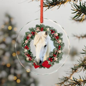 Op de afbeelding: Een cirkelvormig glazen ornament met een wit paardenhoofd in het midden van een feestelijke krans. De krans is versierd met groene dennennaalden, rode bessen, kleine witte sterren en zuurstokken. Een rode strik bevindt zich aan de onderkant.