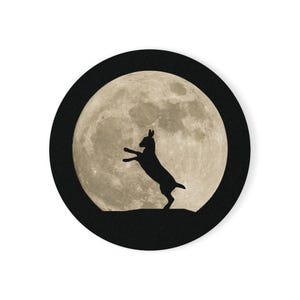 Op de afbeelding: Een ronde sticker met een zwart silhouet van een konijn dat op zijn achterpoten staat tegen een gedetailleerde, getextureerde beige volle maan. Het gehele cirkelvormige ontwerp is omlijst door een effen zwarte rand voor contrast.
