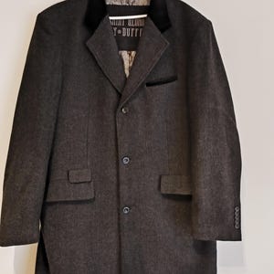 Puede incluir: Un abrigo gris oscuro con cuello y ribetes de terciopelo negro. El abrigo tiene cierre de tres botones, dos bolsillos delanteros y botones en las mangas. La marca "Saint George & Duffy" es visible.