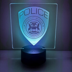 May include: A clear, square LED night light with a police badge design. The badge features the text "POLICE" and "GARDEN CITY MICHIGAN" with a deer emblem. The light emits a blue and teal glow from a black base.