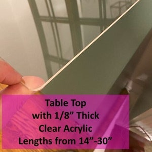 May include: Clear acrylic table top, 1/8" thick, with lengths from 14" to 30". The transparent material reflects the surrounding environment. The image shows a corner of the acrylic sheet, highlighting its clarity and edge detail.