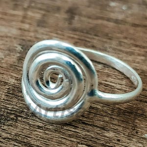 May include: A silver ring with a spiral design. The ring has a coiled, circular pattern at the centre, with a smooth, polished band. The ring is set against a wooden surface, highlighting its metallic shine.