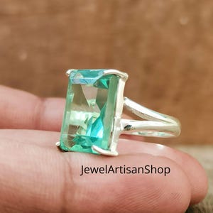 Könnte beinhalten: Ein Silberring mit einem großen, rechteckigen, hellgrünen Edelstein. Der Edelstein ist facettiert, und der Ring teilt sich oben in zwei Teile. Der Text "JewelArtisanShop" ist unten sichtbar.