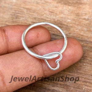 Könnte beinhalten: Ein silberner Ring mit herzförmigem Design. Der Ring wird vor einem Holzuntergrund gehalten. Der Text "JewelArtisanShop" ist unten im Bild zu sehen.