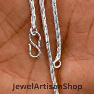Könnte beinhalten: Nahaufnahme einer silbernen Halskette mit Hakenverschluss und kleinem Ringverschluss. Die Kette hat ein gewebtes Design. Der Text "JewelArtisanShop" befindet sich am unteren Rand des Bildes.