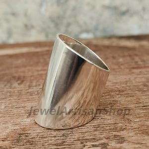 May include: A polished silver ring with a cylindrical design and a slanted top. The ring has a smooth, reflective surface and is set against a wooden background. The text "JewelArtisanShop" is visible on the surface.