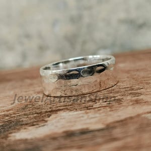 Op de afbeelding: Een zilveren ring met een gehamerde textuur. De ring is breed en heeft een band met een reeks kleine, gelijkmatig verdeelde inkepingen. De ring wordt gepresenteerd op een houten oppervlak.