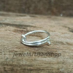Könnte beinhalten: Ein silberner Ring mit einem einzigartigen Design. Der Ring hat ein Doppelband, das sich um den Finger wickelt, mit einem knotenartigen Detail auf der einen Seite und einer kleinen Perle auf der anderen Seite. Der Ring wird auf einer Holzoberfläche gezeigt.