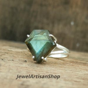 Op de afbeelding: Een zilveren ring met een grote, vliegervormige labradoriet edelsteen met groene en blauwe iridiscentie. De steen wordt op zijn plaats gehouden door vier metalen pootjes op een eenvoudige zilveren band. De tekst JewelArtisanShop is onderaan zichtbaar.