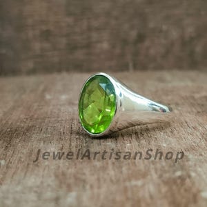 Könnte beinhalten: Ein Silberring mit einem großen, oval geschliffenen, hellgrünen Edelstein. Der Ring hat einen glatten, polierten Ring und eine Lünettenfassung. Der Text "JewelArtisanShop" ist sichtbar. Der Edelstein ist leuchtend grün.