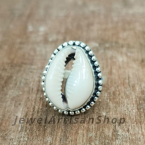 Puede incluir: Un anillo de plata con una concha de cauri blanca como pieza central. La concha está rodeada por un borde plateado con cuentas. El anillo se muestra sobre una superficie de madera, con el texto "Jewel Artisan Shop" visible.