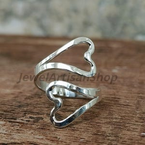 May include: A silver ring featuring two heart-shaped elements. The ring wraps around the finger, with the hearts facing away from each other. The text "JewelArtisanShop" is visible on the ring.